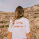 Natural tee with small “be kind” text on front chest and bold “love yourself” text across back
