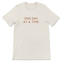 Neutral toned tee with take it one day at a time printed on back, styled casually for an everyday look