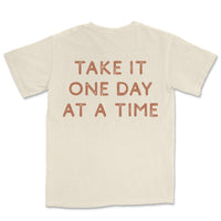 Neutral toned tee with take it one day at a time printed on back, styled casually for an everyday look