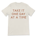 Neutral toned tee with take it one day at a time printed on back, styled casually for an everyday look