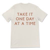 Neutral toned tee with take it one day at a time printed on back, styled casually for an everyday look